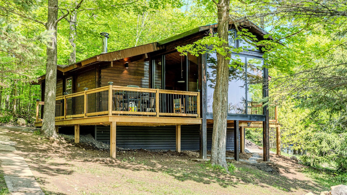 A cottage is nestled in a lush green setting, featuring a wooden deck surrounded by trees. Large windows reflect natural light, providing views of the outdoor landscape. The structure rests on piers, enhancing its picturesque height above the ground.