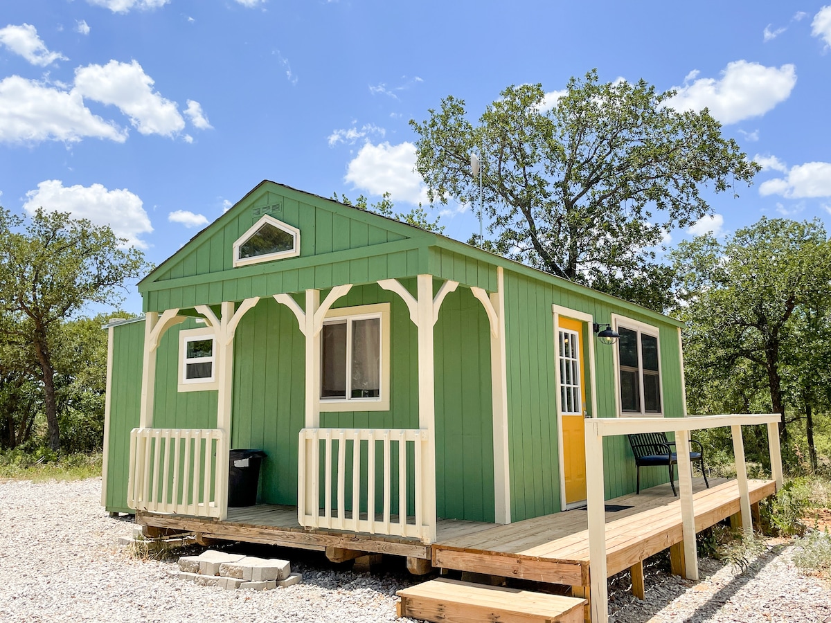 A vibrant green tiny home is shown surrounded by grassy areas and scattered trees. The structure features a bright yellow door and large windows, allowing natural light to flood the space. The outdoor deck includes a small seating area, enhancing the welcoming curb appeal.