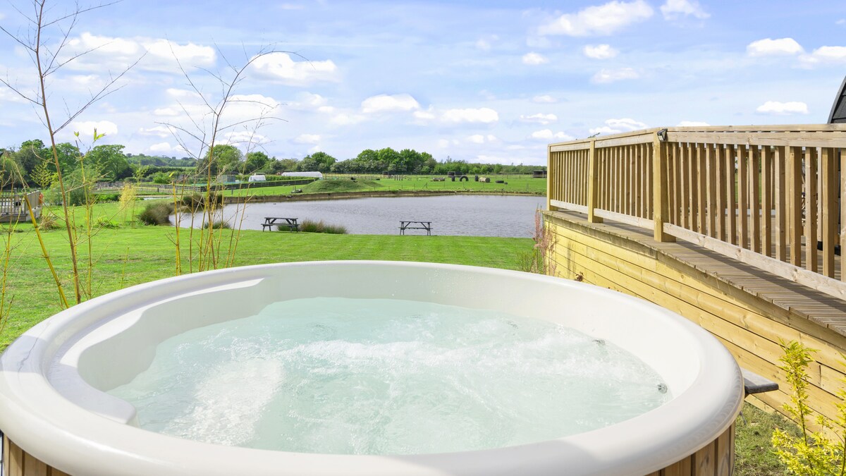 A private wood-fired hot tub is positioned on a wooden patio, surrounded by verdant grass. The scenic view encompasses a serene lake, with picnic tables located on the opposite shore, under a clear blue sky dotted with clouds.