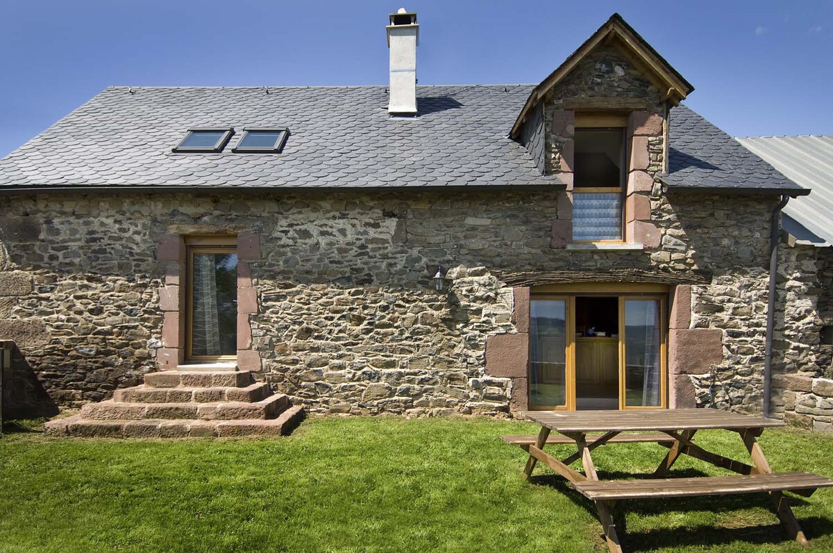 The exterior of the stone cottage features a sloped slate roof and large windows, allowing for natural light. Steps lead up to the entrance, while a wooden picnic table is set on the manicured grass, providing a space for outdoor gatherings.