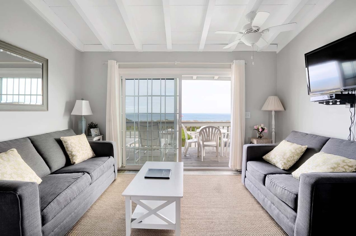 A bright living area features two comfortable gray sofas adorned with decorative pillows. A small white coffee table is centered in front of the sofas. Light flows through a large glass door that opens to a private balcony with outdoor seating, offering scenic views.