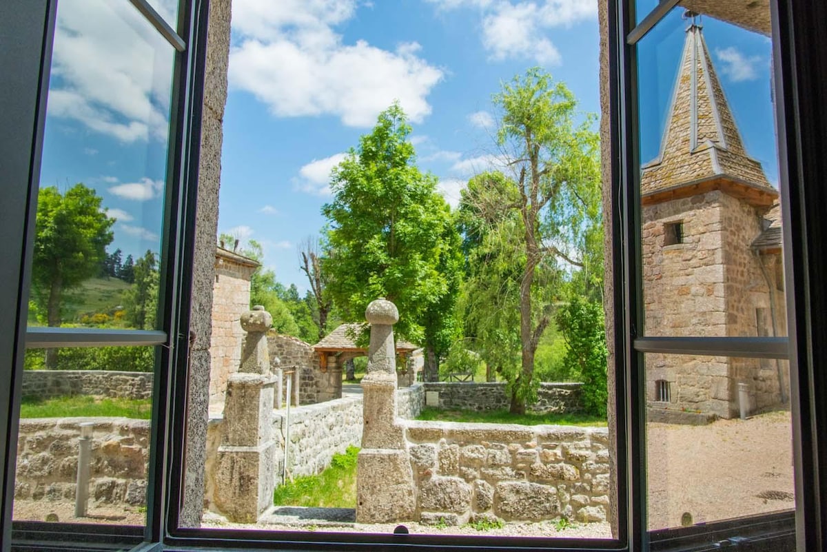 A view through an open window frames a serene outdoor scene, showcasing lush greenery, stone structures, and a blue sky filled with fluffy clouds. The rustic charm of the surroundings is highlighted by the well-maintained stone walls and the distant tower.