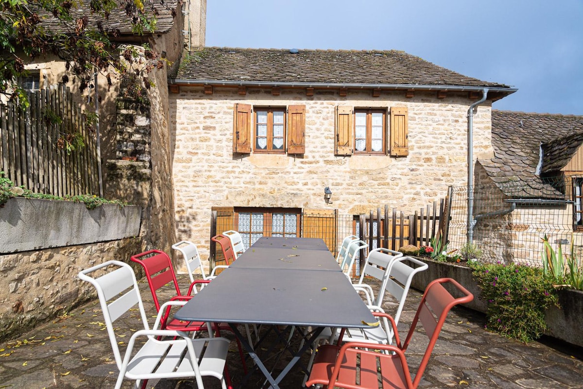 An outdoor dining area features a long table surrounded by a mix of red and white chairs. The rustic stone building serves as a backdrop, displaying wooden shutters on windows. Greenery and stone flooring add a natural touch to the setting.