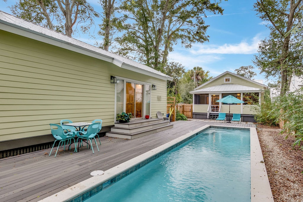 A modern cottage home is shown beside a private outdoor pool. The area features a wooden deck with blue outdoor chairs and a table, alongside the brightly colored pool house. Lush greenery surrounds the pool, contributing to the tranquil atmosphere.