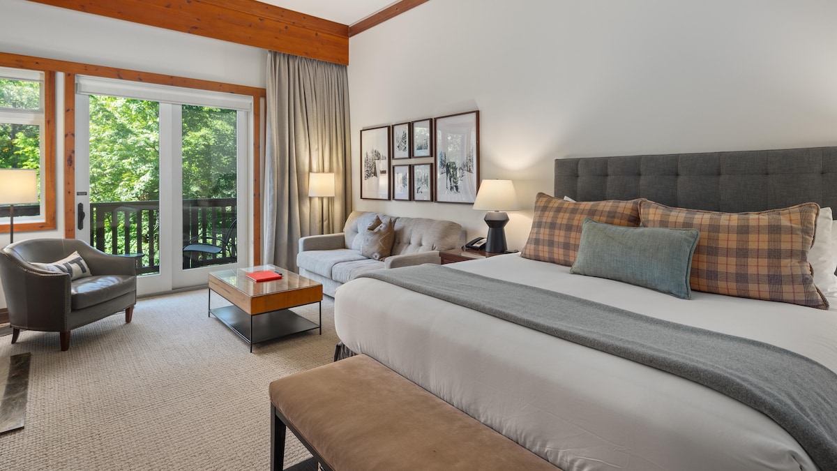 A studio room features a spacious bed with neutral bedding and accent pillows. A comfortable sofa and chair are positioned near large sliding glass doors, offering views of surrounding greenery. Soft lighting is provided by table lamps on either side of the bed.