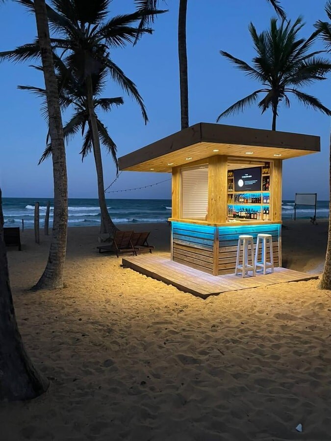 A beachfront bar is illuminated at dusk, featuring a wooden structure with a blue and natural finish. Tall palm trees frame the scene, and waves are visible in the background. Two white bar stools are positioned in front of the bar, inviting guests to relax.