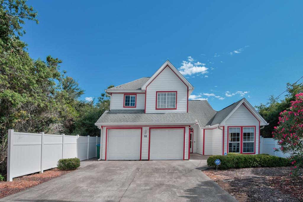 The two-story beach house features a light exterior with red accents, framed by well-maintained greenery and a white privacy fence. A paved driveway leads to the double garage, while large windows allow for ample light. The clear blue sky enhances the bright appearance of the home.