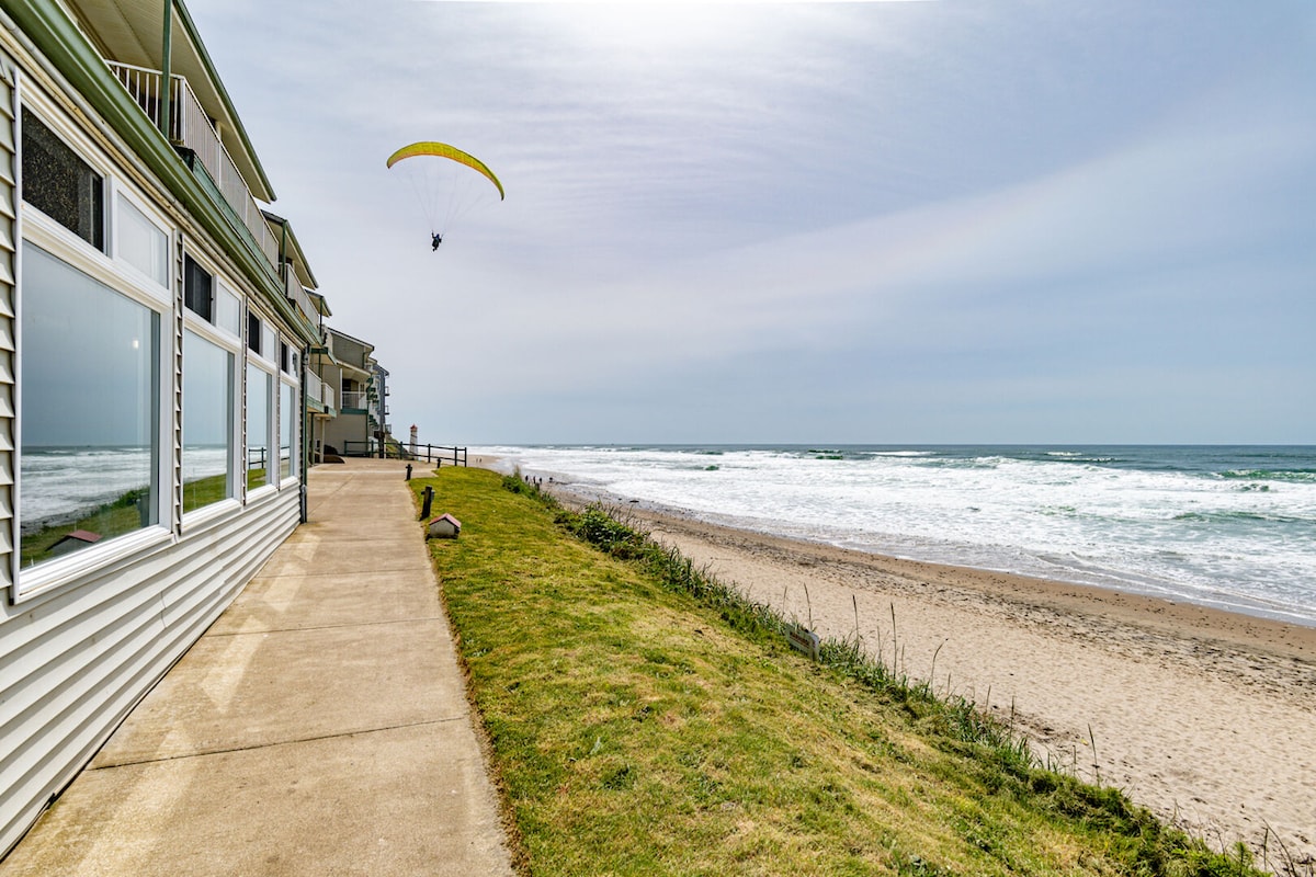 A smooth pathway runs alongside the beach, offering a view of the ocean waves and a colorful yellow parachute in the sky. Nearby, well-maintained grass and a sandy beach create a tranquil coastal setting, with condominium units visible on the left.