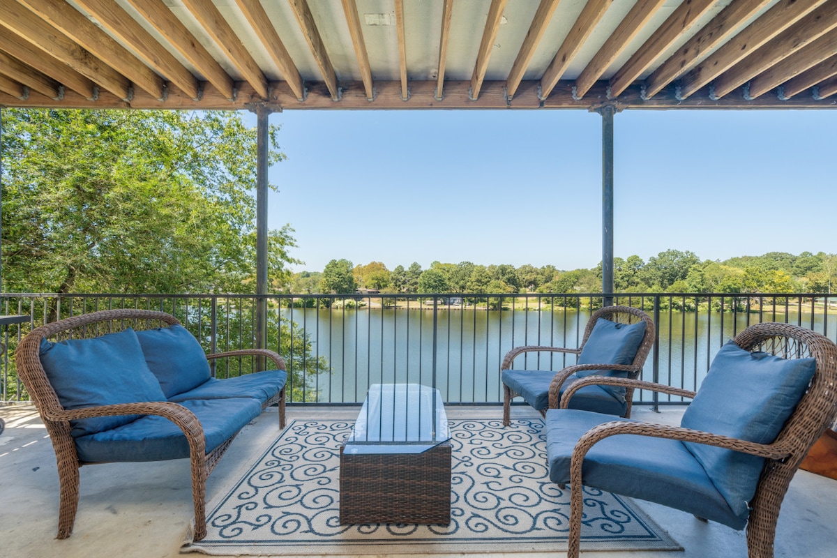 A covered balcony features four outdoor chairs and a glass-top coffee table, all arranged on a decorative area rug. The tranquil view of Lake Hamilton and surrounding greenery is visible through the railing, creating a serene outdoor space.