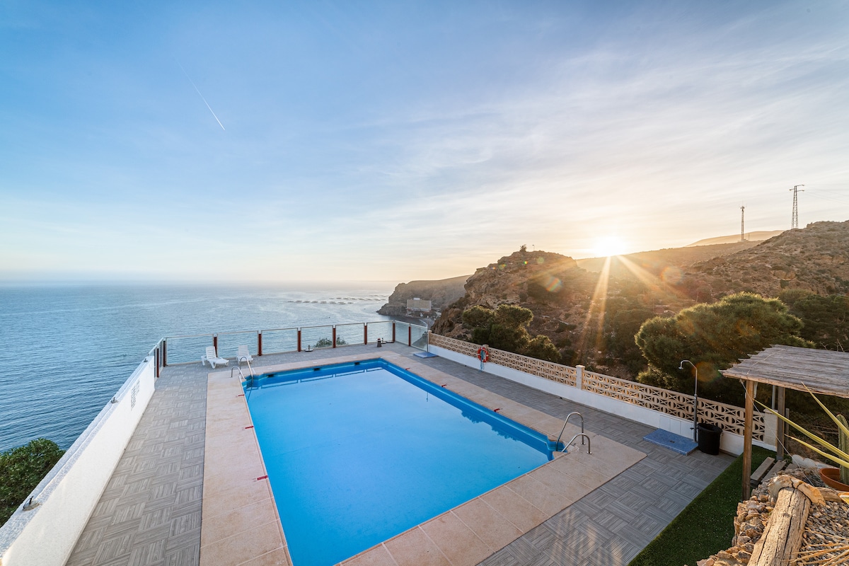 A clear view of a spacious swimming pool is presented, set against a backdrop of the sea and rocky cliffs. The pool area is bordered by a tiled deck, and the horizon features a soft sunset glow.