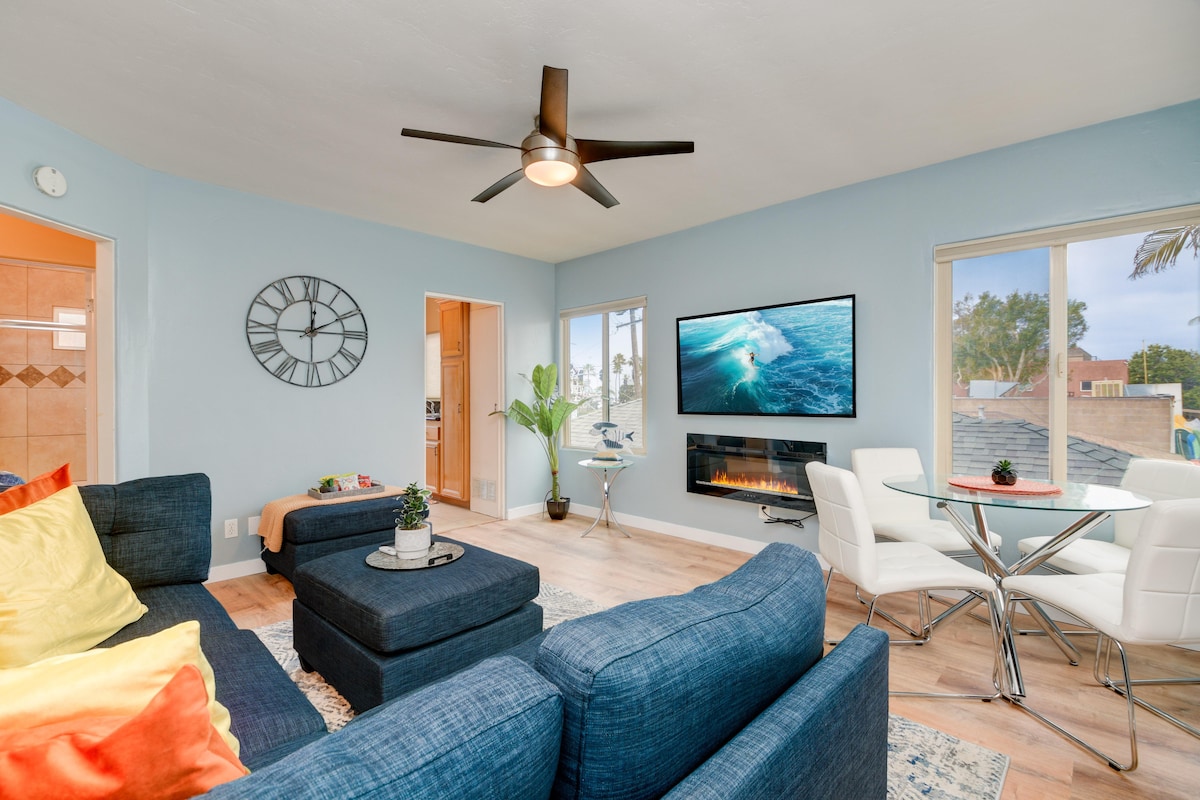A bright living area features a spacious blue sectional sofa adorned with colorful cushions. A flat-screen television is mounted on the wall, complemented by a modern electric fireplace below. Large windows allow natural light to illuminate the room, with a dining table for four nearby.