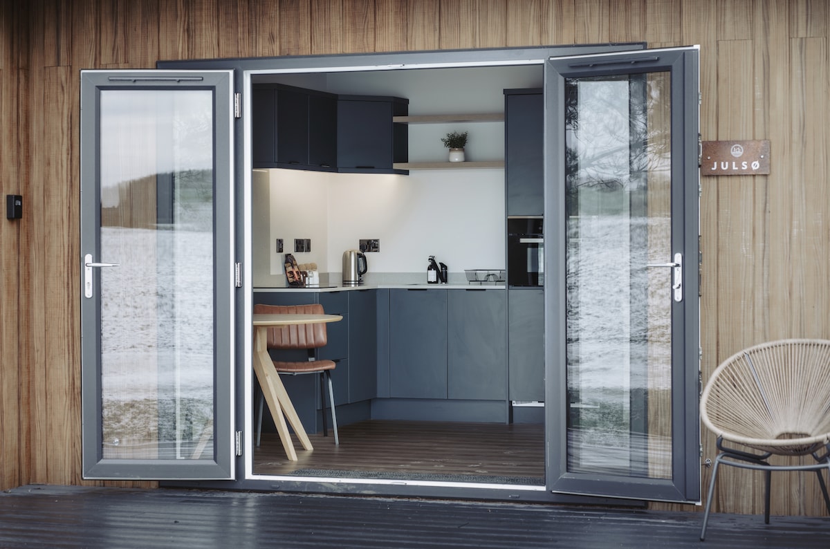 The modern kitchen area is visible through wide-opening glass doors, showcasing sleek cabinetry and kitchen appliances. A wooden dining table and chair are positioned nearby. The wooden deck extends outward, creating a seamless transition between indoor and outdoor spaces.