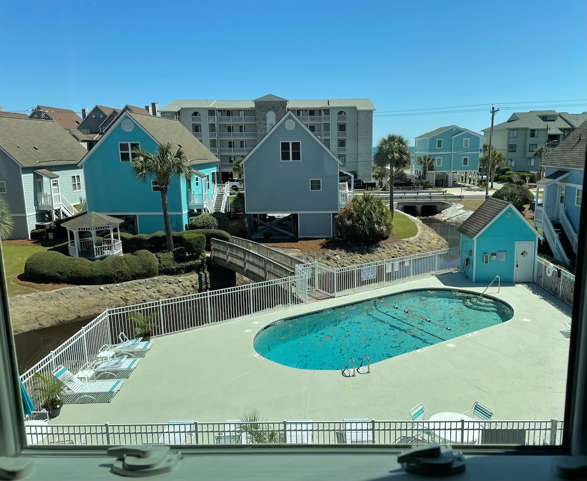 An overhead view captures a community pool, surrounded by a white fence. Colorful houses line the background, with palm trees adding greenery. Sunlight brightens the scene, creating a relaxed atmosphere. The pool area features lounge chairs for enjoying the outdoors.
