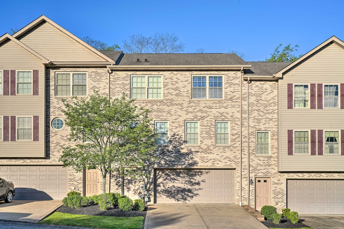 The exterior view of the multi-level townhome is presented, highlighting a beige brick facade and multiple windows. Two garage doors are visible, accompanied by well-maintained landscaping, including trees and shrubs. The clear blue sky provides a bright backdrop.