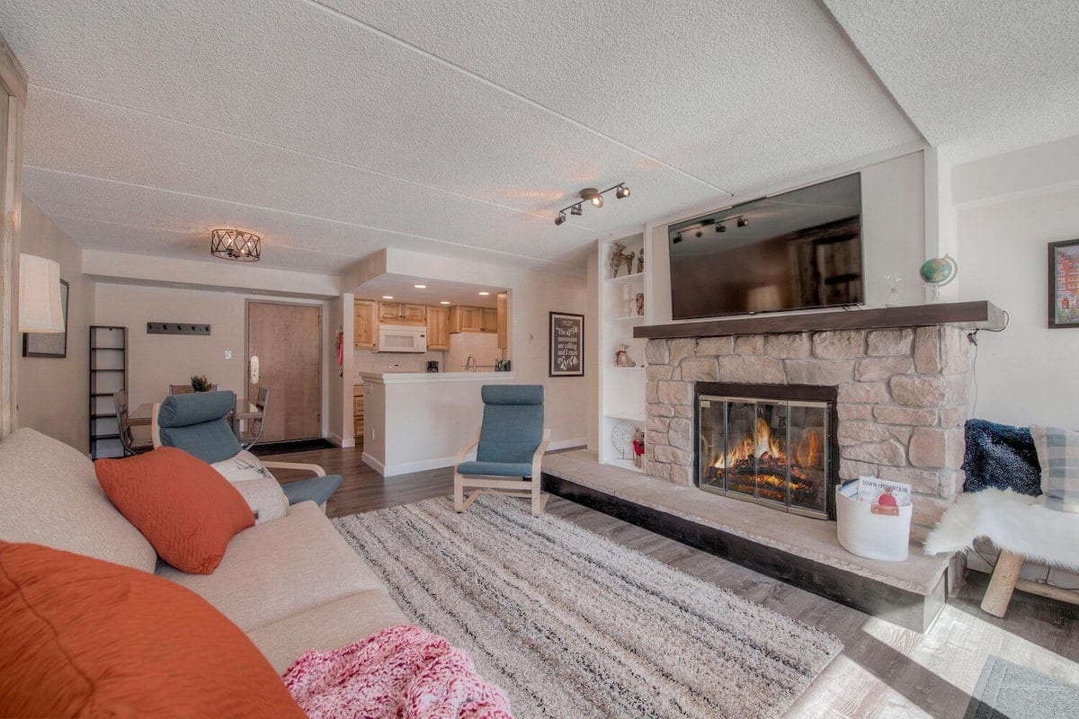 A spacious living area features a stone fireplace with a cozy fire, surrounded by a light-colored sofa adorned with vibrant cushions. Two comfortable chairs offer additional seating. A large television is mounted above the fireplace, while a kitchen area is visible in the background.