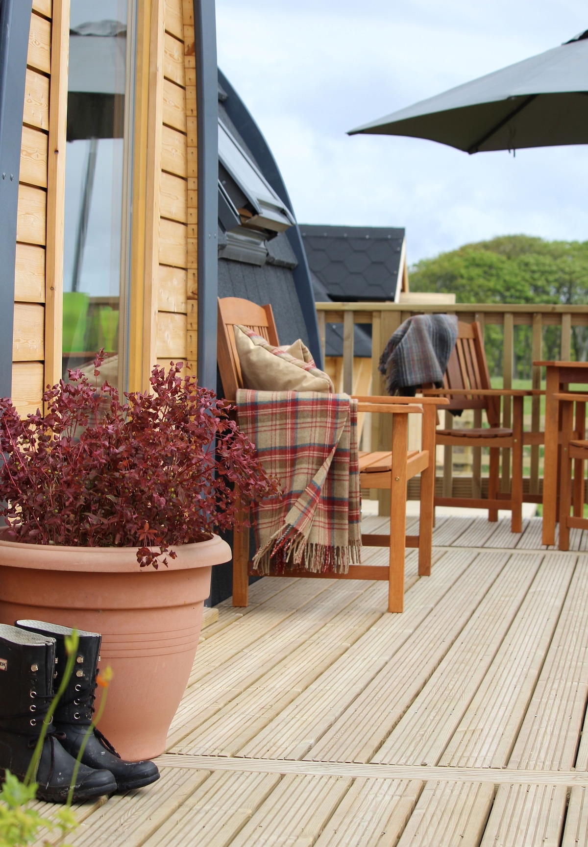 A spacious deck is shown, featuring wooden chairs around a table under a large umbrella. A terracotta plant pot with purple foliage adds a touch of greenery, while a folded plaid blanket drapes over one of the chairs.