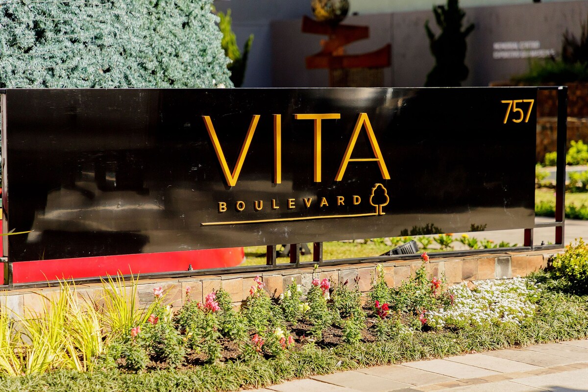 The entrance sign of Vita Boulevard is prominently displayed, featuring golden lettering against a black background. Surrounding the sign is a well-maintained landscape with colorful flowers and greenery, contributing to the inviting atmosphere of the complex.