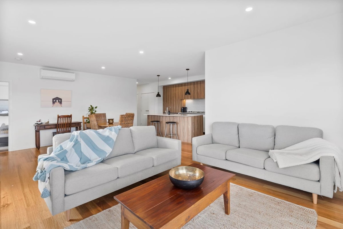 A light-filled living area features two gray sofas positioned around a wooden coffee table. Soft, neutral tones enhance the spaciousness, while a dining table is visible in the background, complementing the open-plan layout. The kitchen area boasts modern cabinetry and pendant lighting.