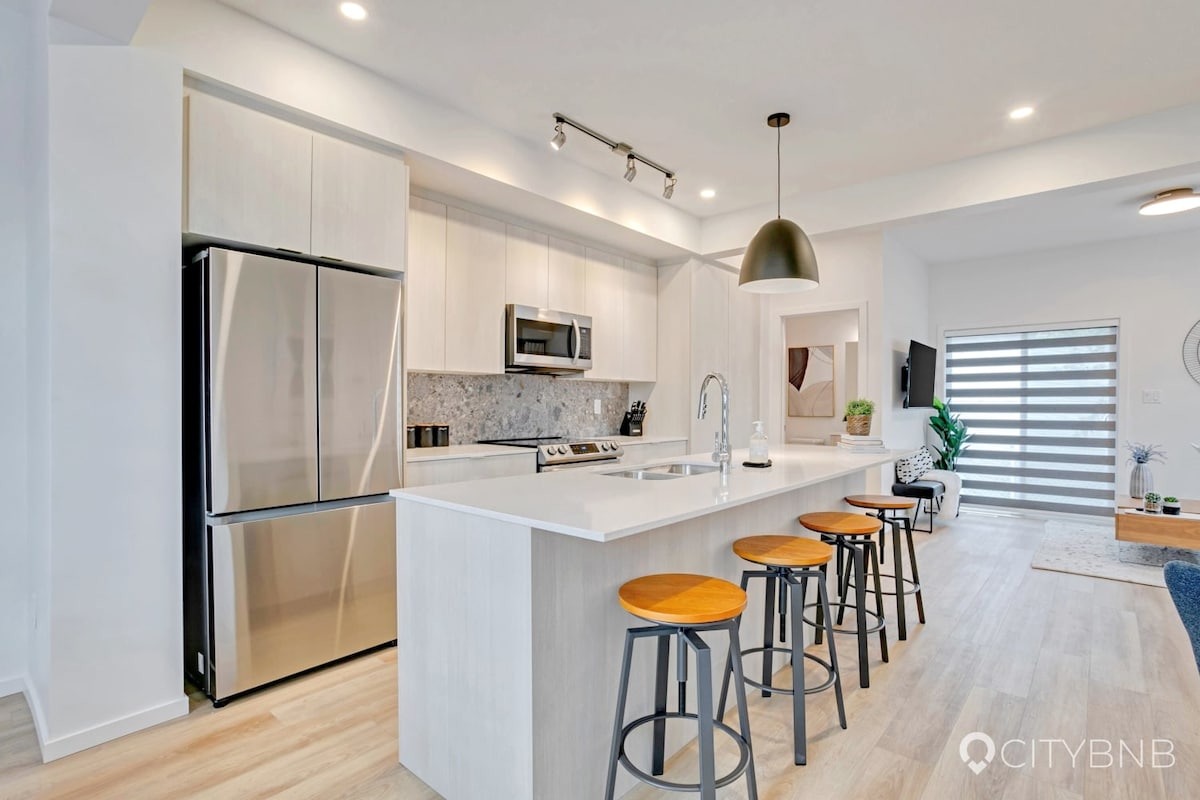 A modern kitchen is highlighted by a large island with four bar stools, sleek cabinetry, and stainless steel appliances. Soft lighting enhances the workspaces, while the textured backsplash adds a touch of elegance. Natural light streams in through a nearby window, illuminating the open layout.