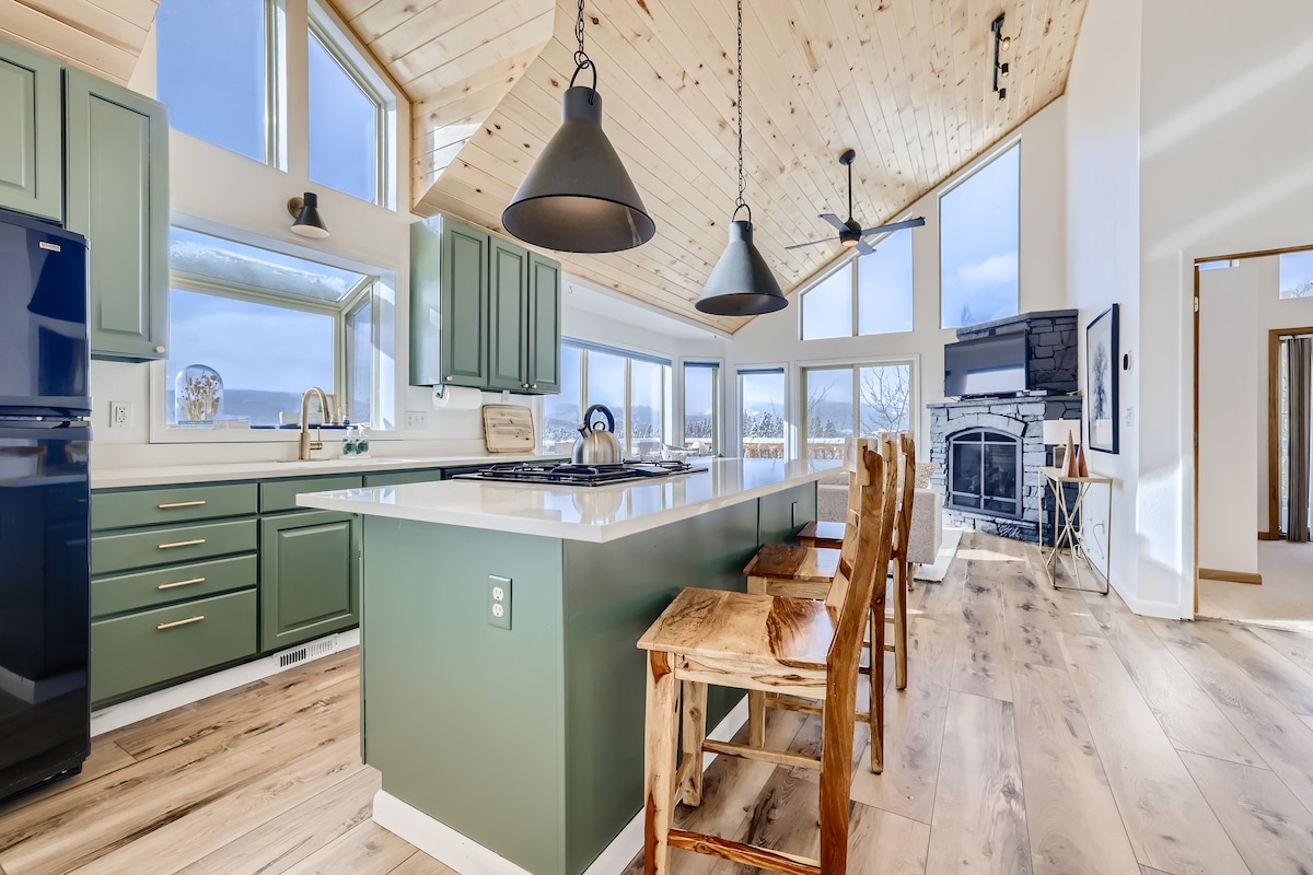 A modern kitchen features green cabinetry complemented by light-colored countertops. Natural light floods in through large windows. High ceilings are accented by wooden beams. Bar seating is provided by several wooden stools arranged along the countertop, with a gas fireplace visible in the adjacent living area.