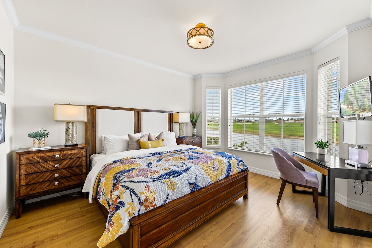 A well-appointed bedroom features a spacious bed adorned with a colorful blanket. A wooden dresser stands alongside the bed, and a sleek desk is positioned by a large set of windows, offering views of the surrounding landscape and allowing natural light to fill the room.