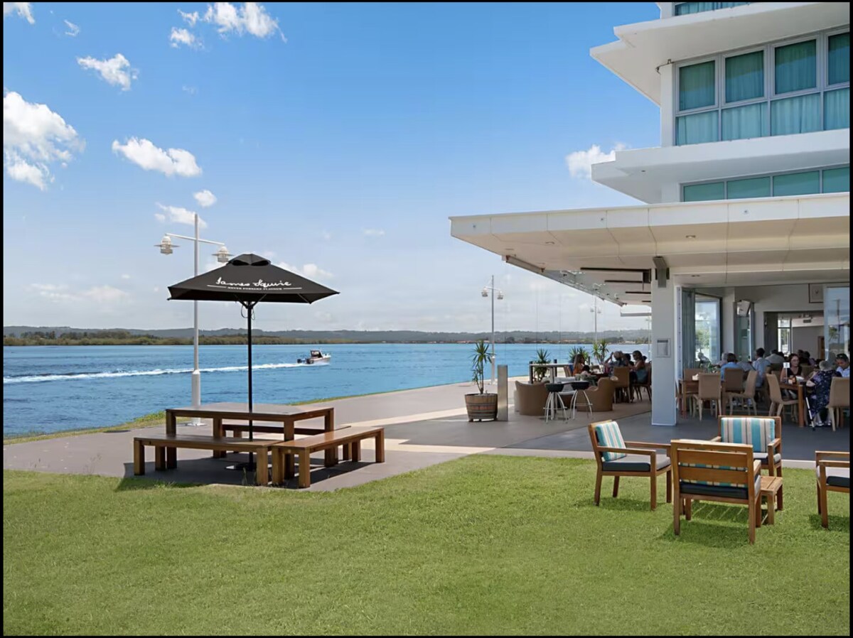 An outdoor dining area features wooden tables and chairs positioned on a grassy lawn. An umbrella provides shade to some seating. The Richmond River is visible in the background, with a boat navigating the water under a clear blue sky.