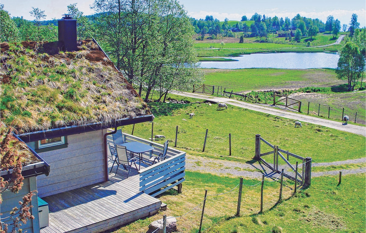 A cottage with a green, grass-covered roof overlooks a serene lake. A wooden terrace with outdoor seating is visible, surrounded by lush greenery. A gravel path leads through the scenic landscape, which features open fields and a fence in the distance.