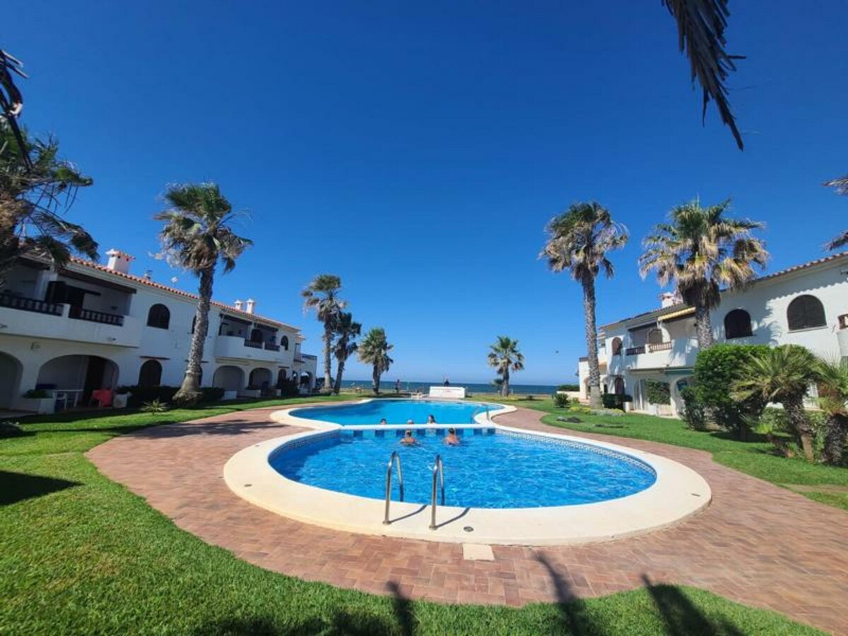 A communal pool is framed by well-maintained gardens and palm trees. The pool design features a curved shape, with gentle steps leading into the clear blue water. The surrounding lawn is neatly kept, providing a relaxing atmosphere for guests.