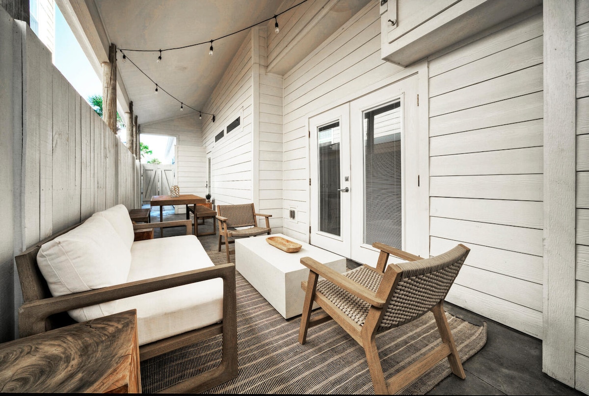 A covered patio area is furnished with a large seating arrangement, featuring neutral-colored cushions and wooden accents. A central coffee table and a small side table provide functional space. Soft lighting is strung across the ceiling, enhancing the welcoming ambiance.