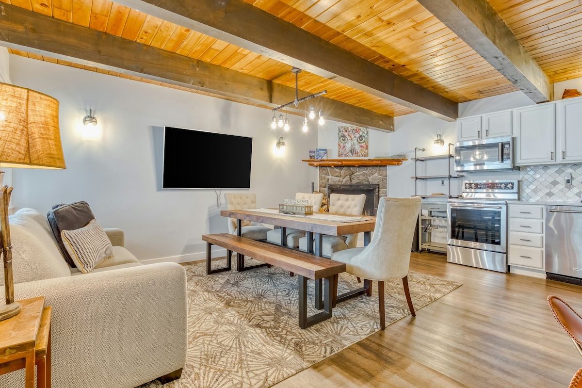 A spacious living area features exposed wooden beams overhead and a cozy sofa alongside a light-colored rug. A large wall-mounted TV is positioned above a stone fireplace. A modern dining table with upholstered chairs is set adjacent to a well-equipped kitchen, highlighted by stainless steel appliances.