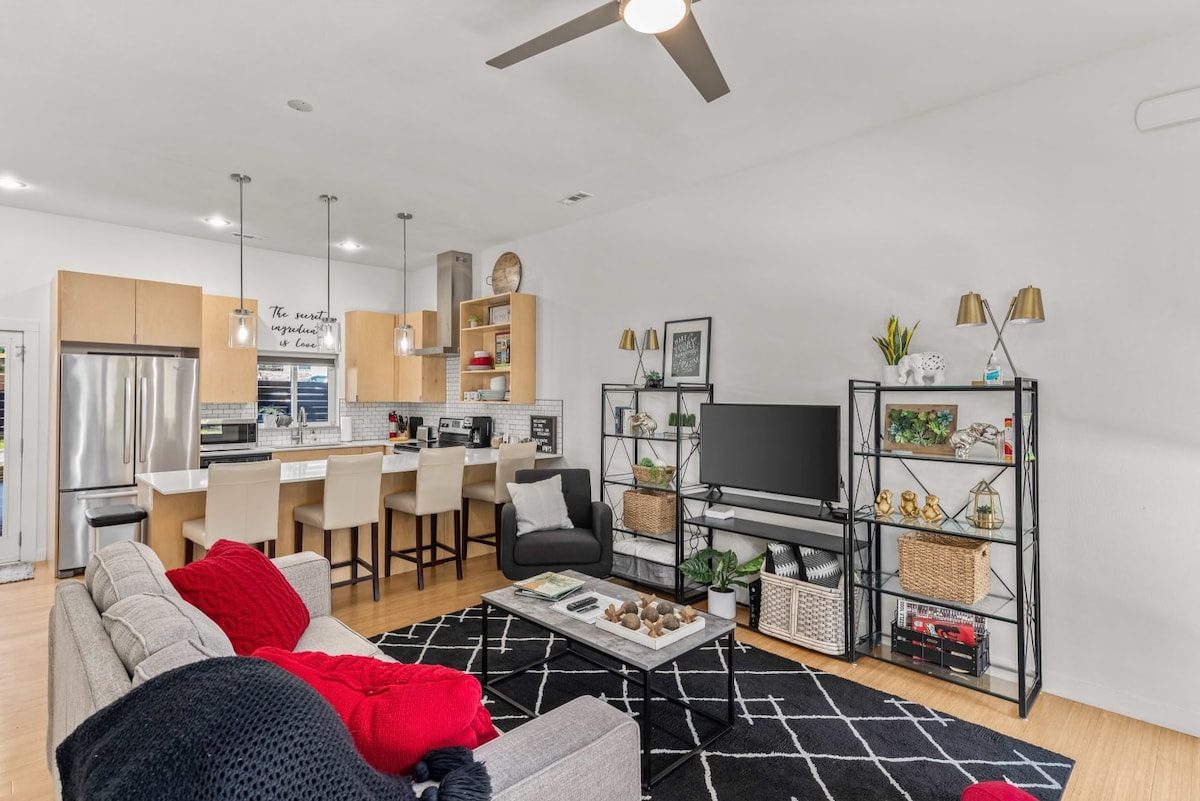 The open-concept living area features a modern design with a spacious gray sofa adorned with red throws. A large TV is positioned on a black shelving unit, complemented by decorative plants and baskets. The kitchen area showcases light-colored cabinetry and stainless-steel appliances, with a dining bar featuring stools.