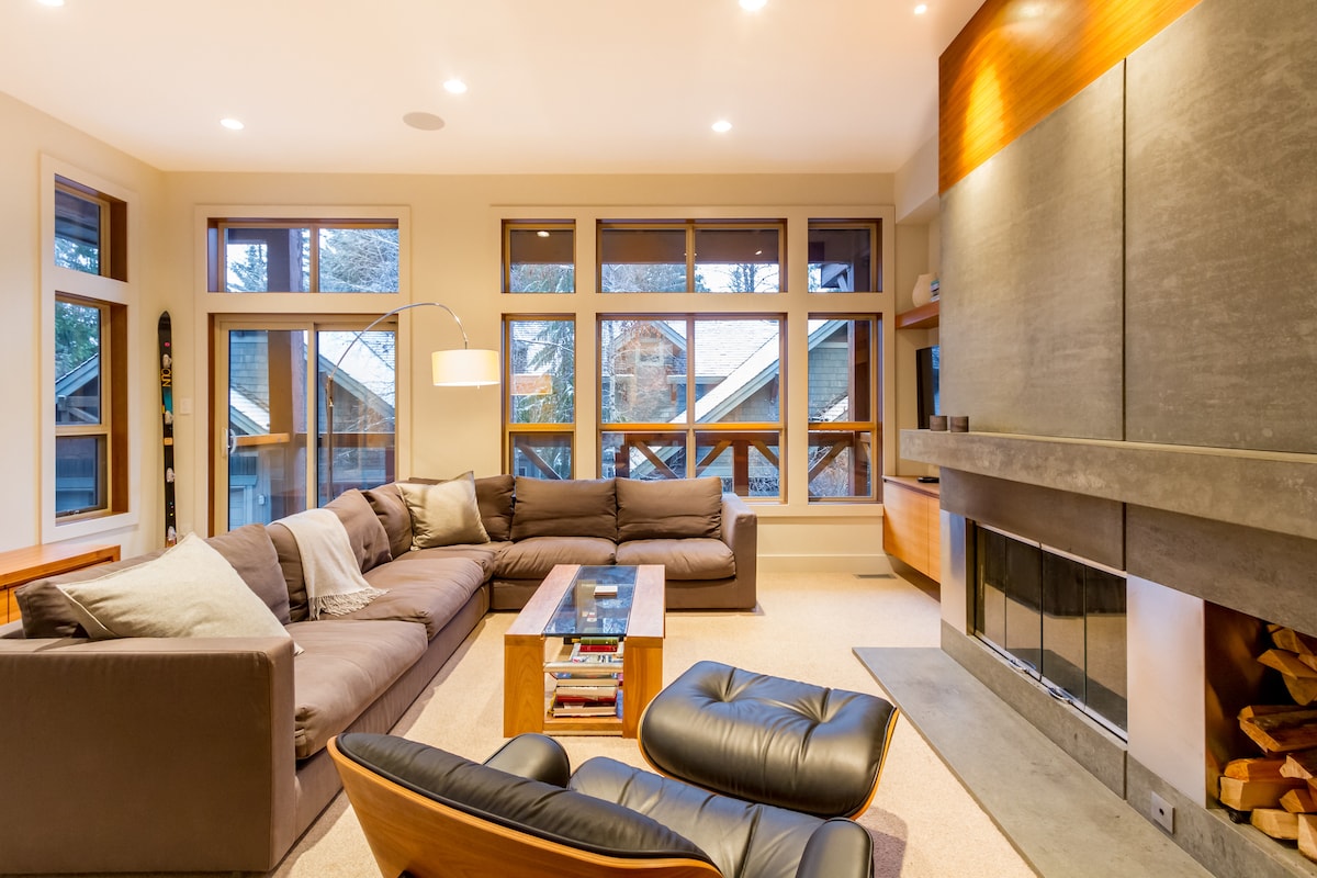 A spacious living area is accented by large windows, allowing natural light to fill the room. A sectional sofa and a sleek lounge chair provide comfortable seating. A stone fireplace adds a modern touch, while a wooden coffee table offers a central gathering space.