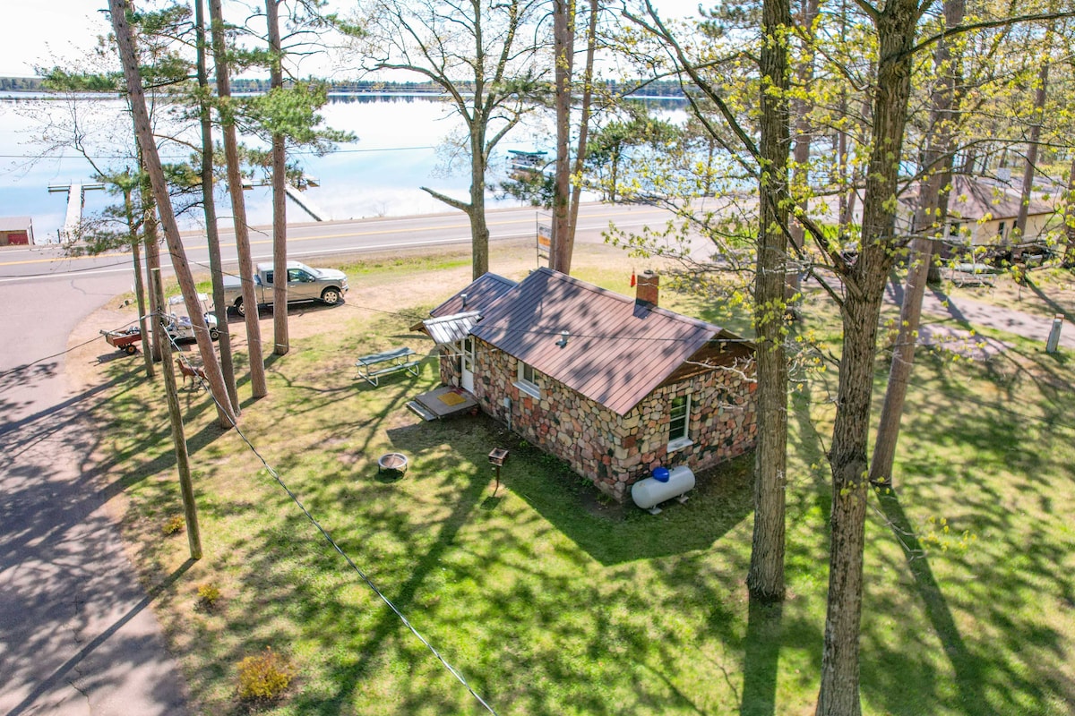 The stone cottage is surrounded by tall trees and lush grass, offering a serene outdoor space. A gravel driveway is visible, along with a vehicle parked nearby. The expansive lake can be seen in the background, providing a scenic view.