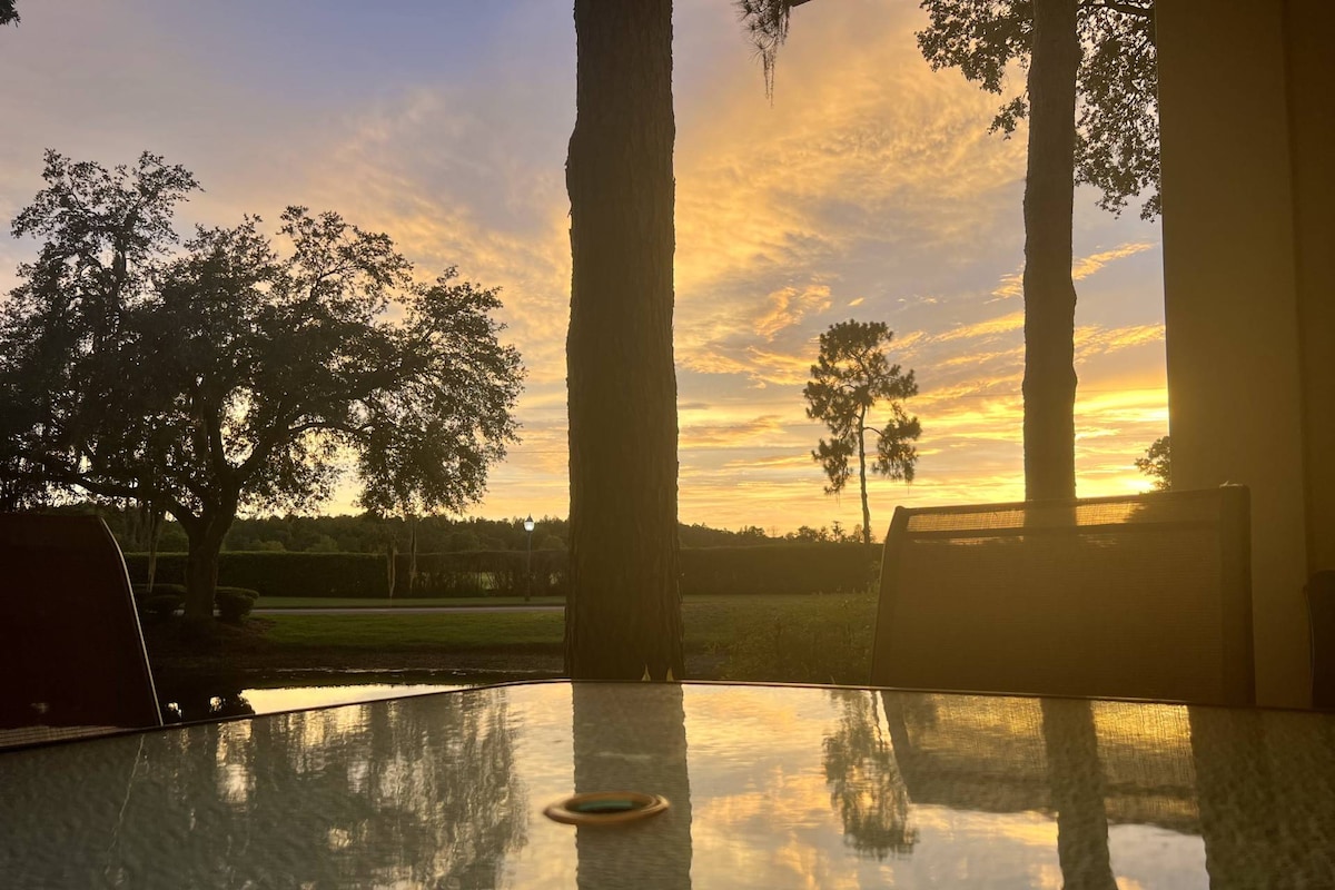 A tranquil outdoor patio scene showcases a table reflecting the warm colors of a sunset. Silhouetted trees and a peaceful landscape are visible in the background, creating a serene atmosphere perfect for relaxation.