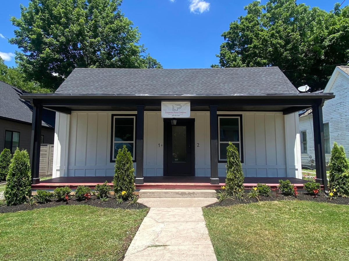 A charming single-story structure is presented, featuring a clean façade with a dark roof and white siding. The front porch is bordered by neatly trimmed shrubs and vibrant flowers. A concrete pathway leads to the entry, enhancing accessibility and inviting guests.