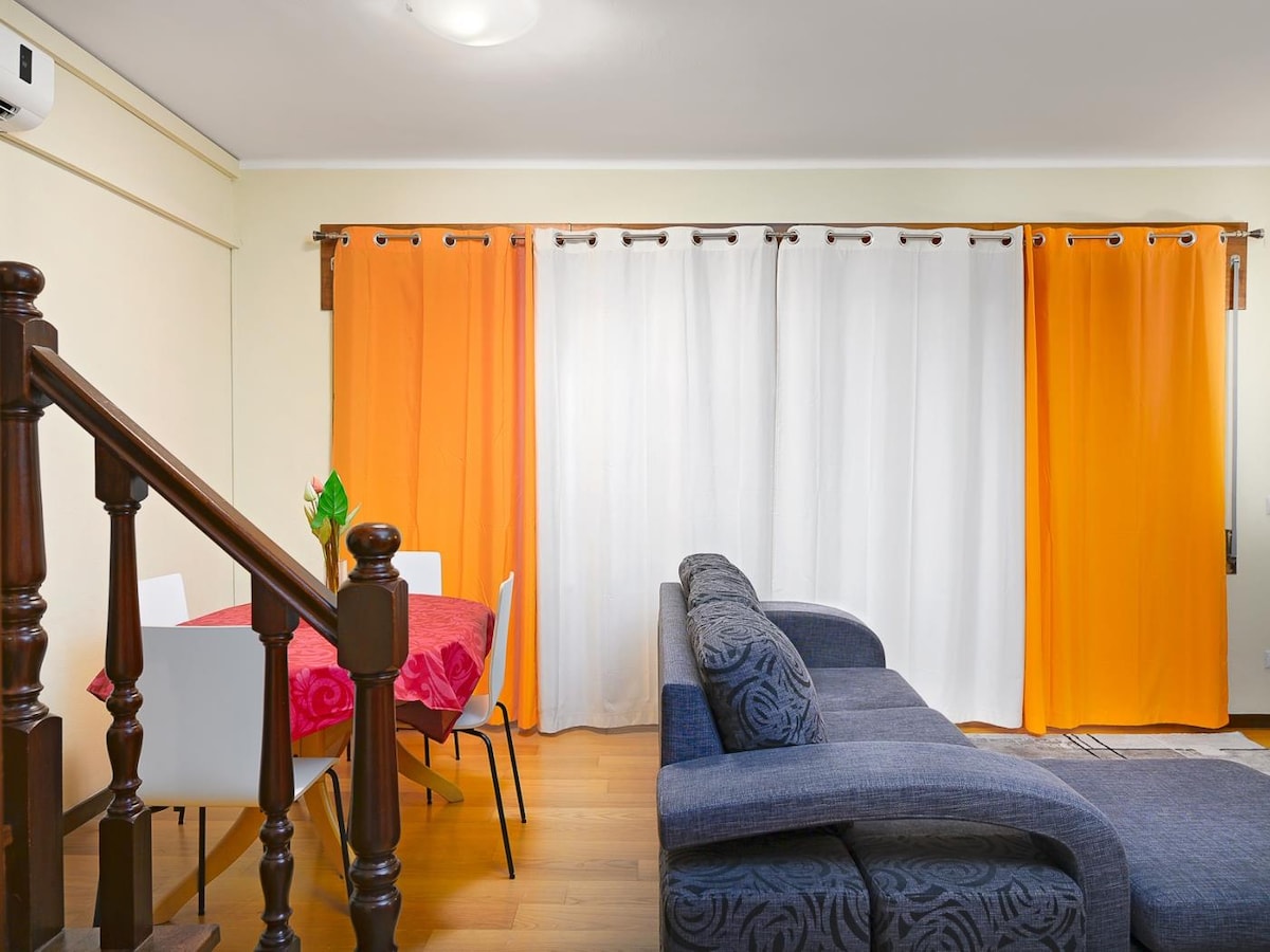 A cozy living area is positioned beside a dining space. Bright orange and white curtains frame a large window, allowing ample natural light. A comfortable dark sofa is complemented by a simple dining table set for four, creating an inviting atmosphere.