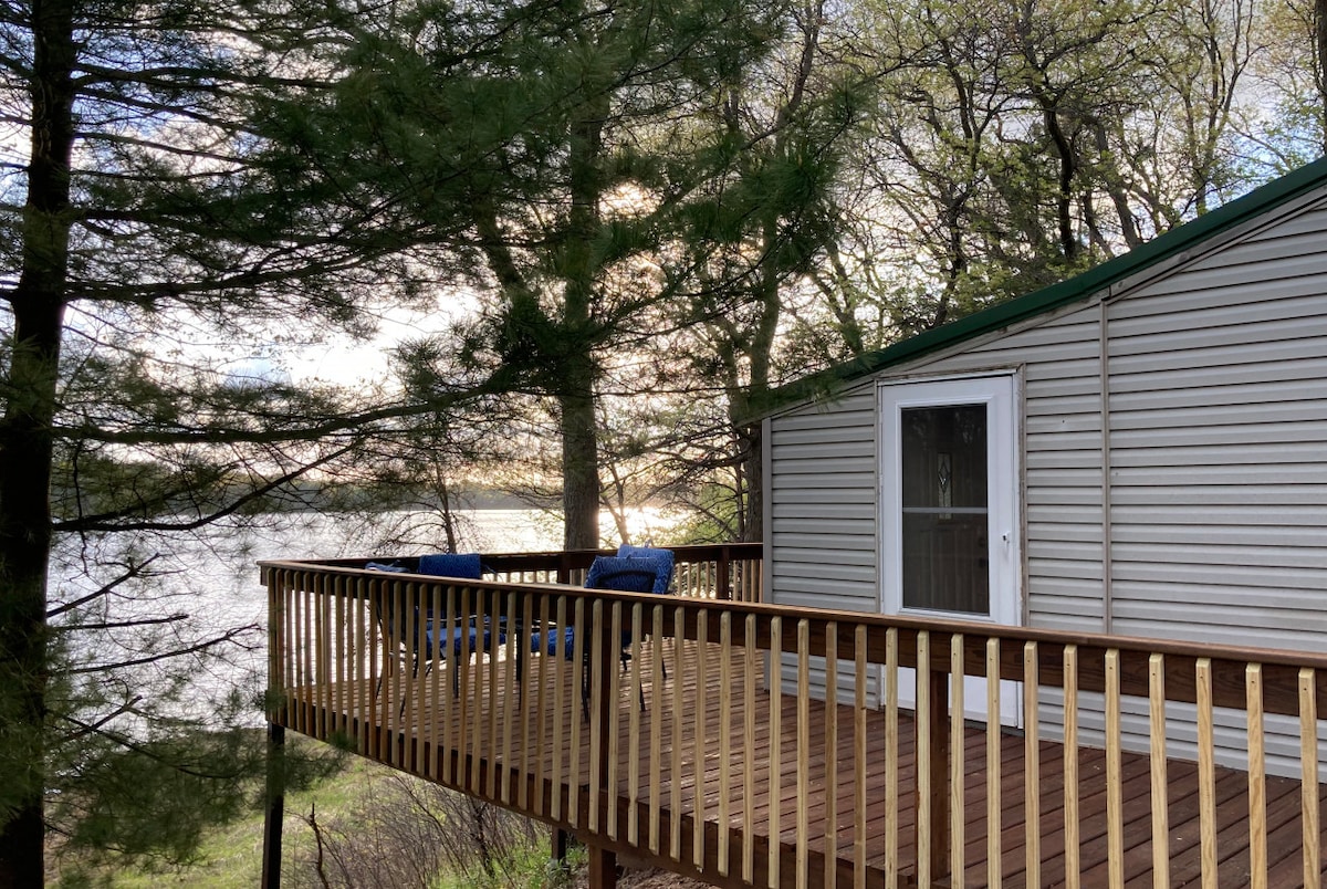 A large wrap-around deck is visible, featuring two blue chairs positioned for a view of the water. Sunlight filters through the trees, creating dappled patterns on the deck's wooden surface, while the calm water reflects the soft hues of the sky.