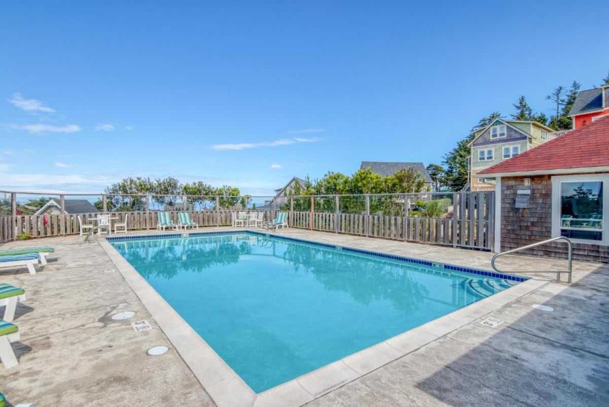 A community pool is displayed, surrounded by a wooden fence and sun loungers. The clear water reflects the sky, while beach houses are visible in the background. The space offers a relaxed setting for guests to enjoy the outdoors.