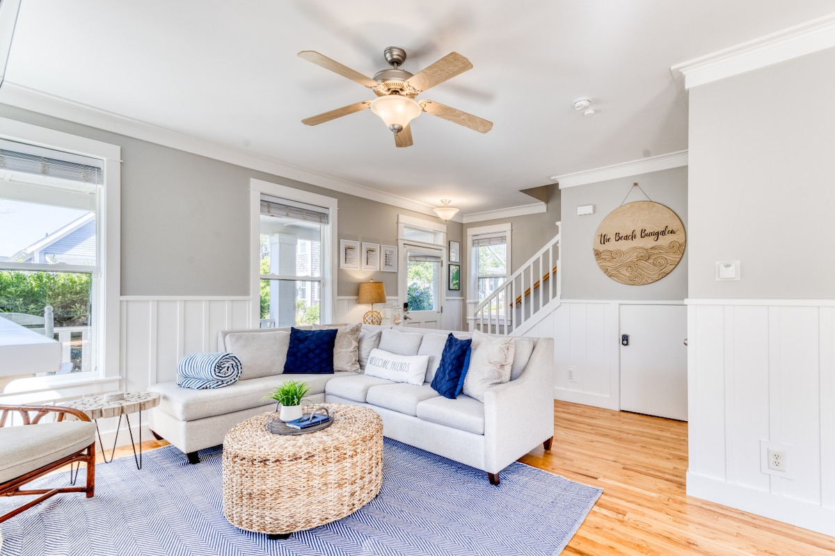 A light-filled living area features a comfortable gray sofa adorned with blue pillows, positioned next to a woven coffee table. Natural light enters through multiple windows, illuminating the space. A ceiling fan and decorative wall sign enhance the home's welcoming ambiance.