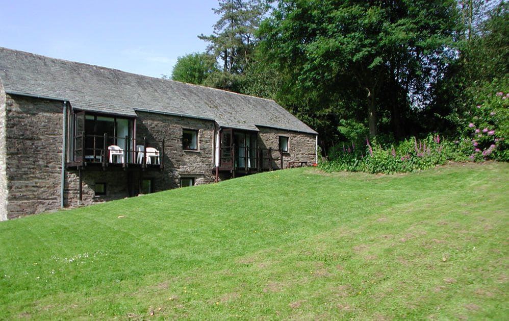 The exterior of Grange Cottage is shown, featuring a stone façade with large glass doors leading to balconies. A grassy area surrounds the cottage, adorned with trees and shrubs. A clear blue sky provides a bright backdrop to the peaceful setting.