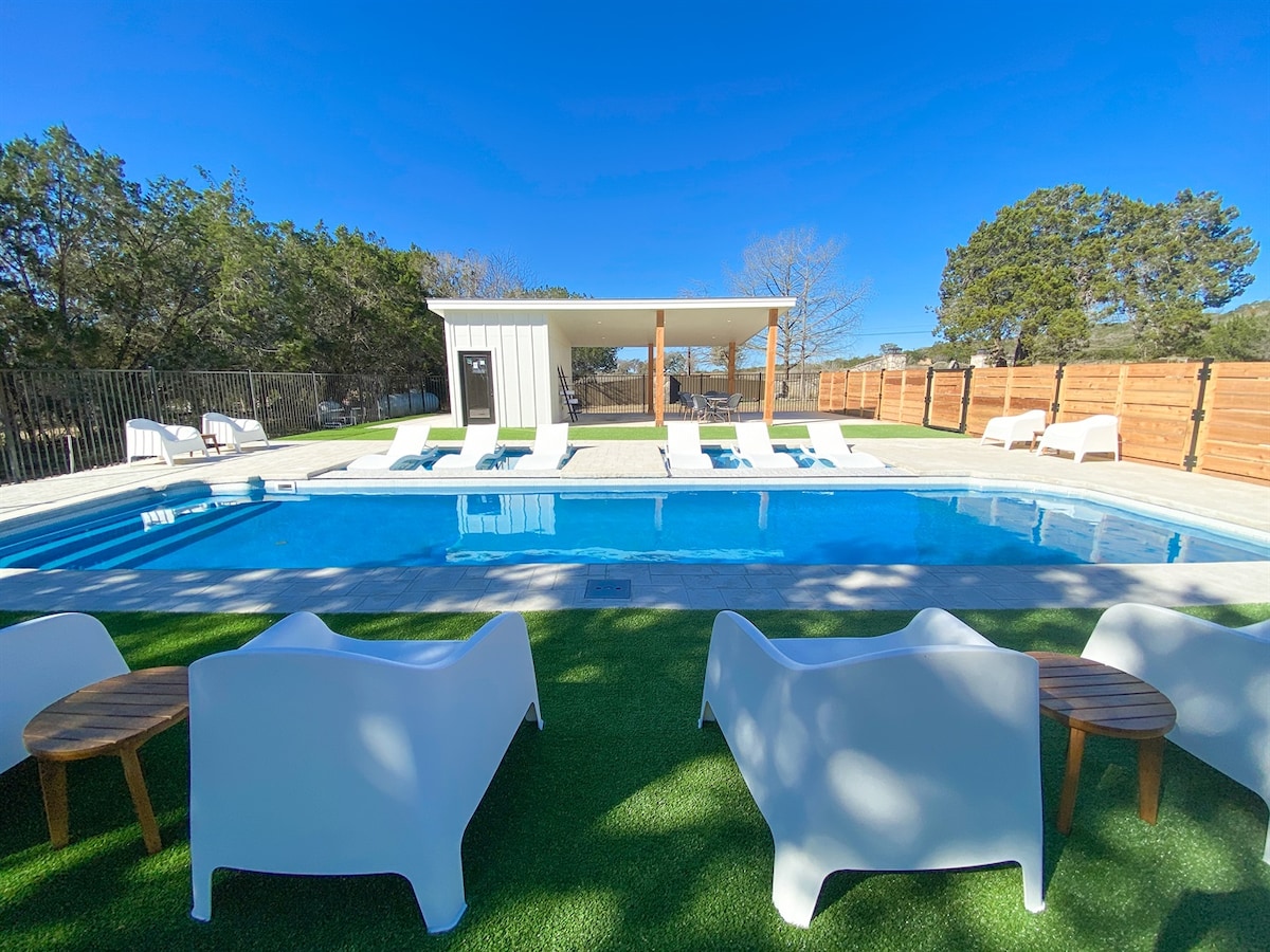 An outdoor resort pool area features a large, shimmering pool, surrounded by lounge chairs and a shaded cabana. A manicured lawn provides a comfortable setting for relaxation, with natural scenery of trees in the background and clear blue skies above.