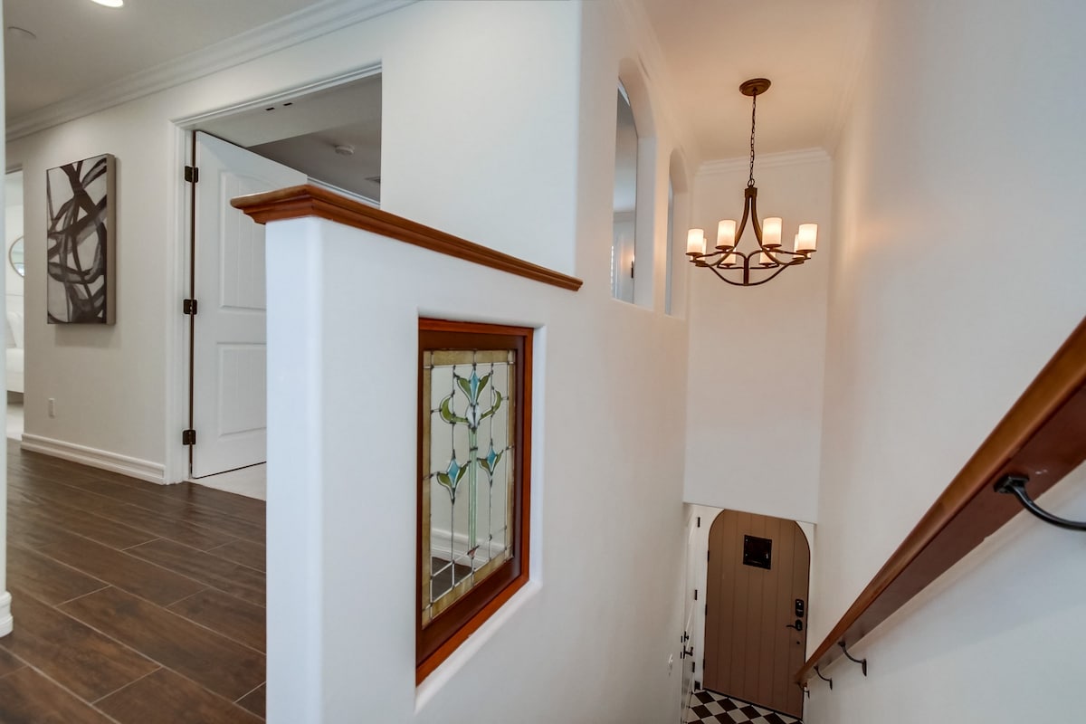 A well-lit staircase leads to the second level, featuring a decorative stained glass window and a stylish light fixture overhead. The walls are painted white, and a patterned black-and-white tile floor is visible at the entrance.