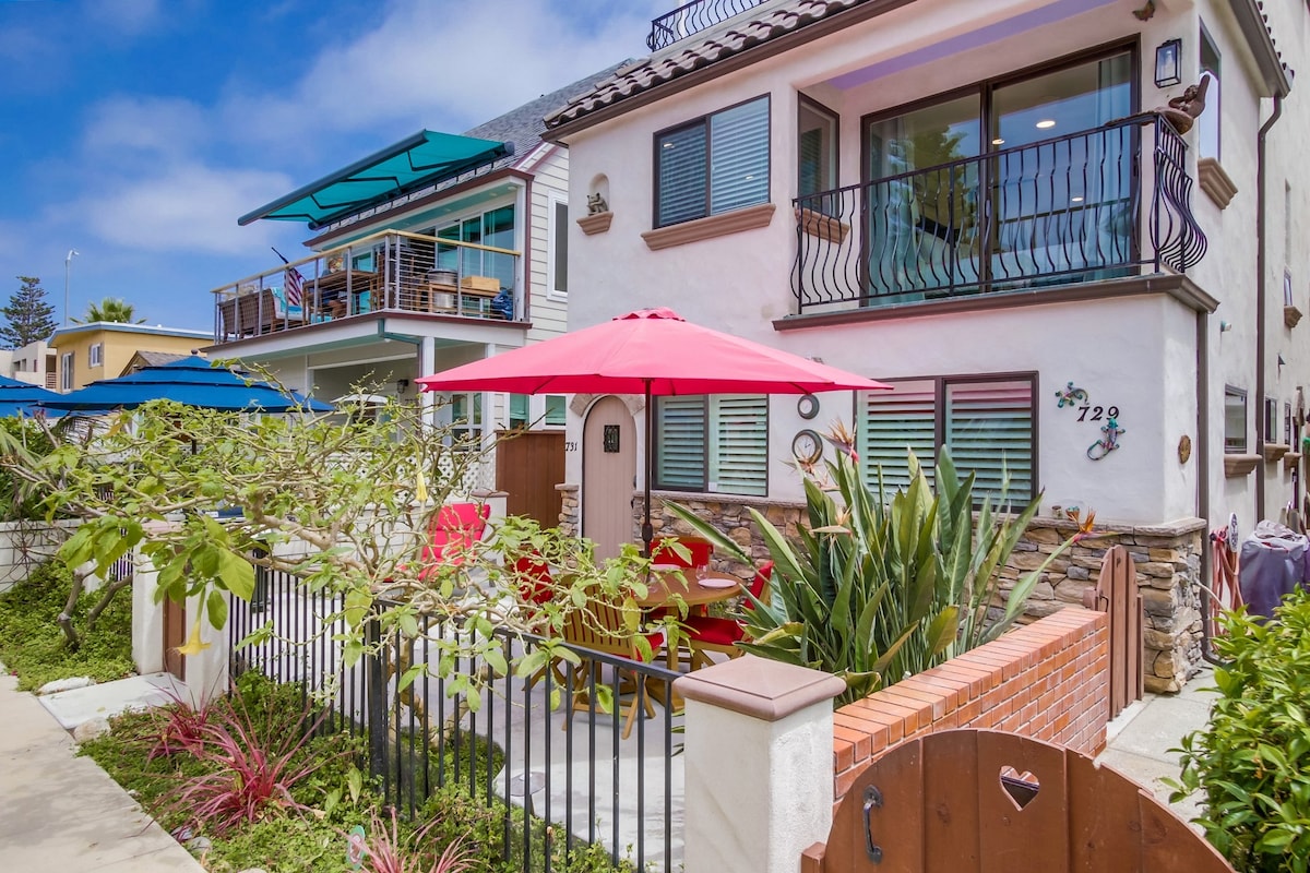 The front patio features a vibrant red umbrella providing shade over a round dining table with colorful chairs. Lush greenery and tropical plants adorn the landscaped area, while a charming brick wall and gated entrance frame the property. The two-story duplex home is seen in the background.