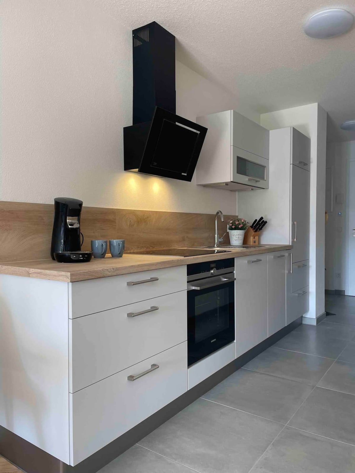 A modern kitchen displays sleek cabinetry and a wood countertop. Equipped with a coffee maker, oven, and an integrated stovetop, it is designed for culinary pursuits. Soft lighting enhances the space, while an adjacent wall houses a built-in microwave and refrigerator.