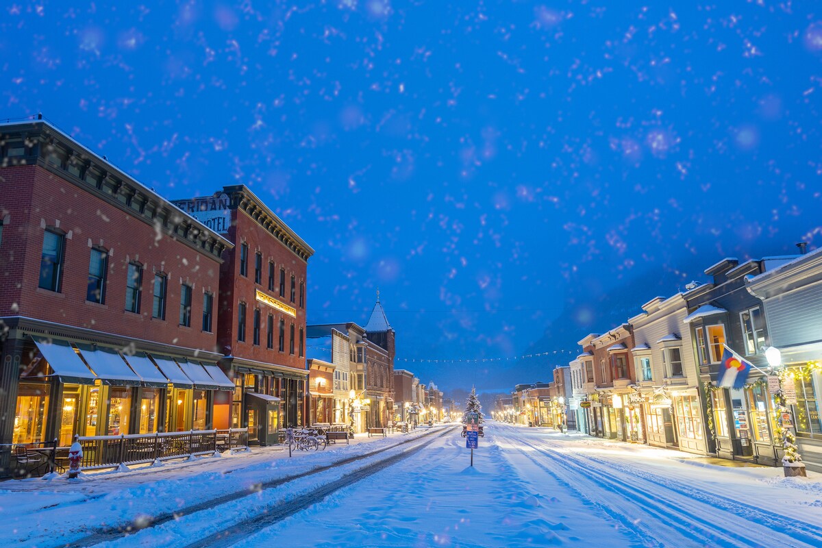 A snow-covered main street is illuminated by soft lights, showcasing a line of historic buildings on either side. The scene reflects a peaceful winter atmosphere, with a gentle snowfall adding a serene touch to the landscape.