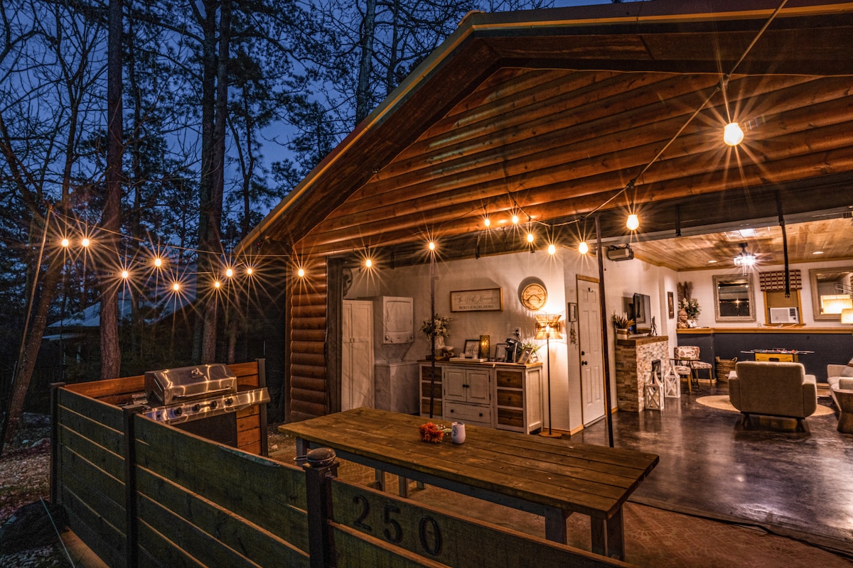An outdoor patio area is illuminated by string lights, featuring a large wooden picnic table and a propane grill. The open cabin interior is visible, showcasing a cozy living space with modern decor and soft lighting, surrounded by a wooded setting.