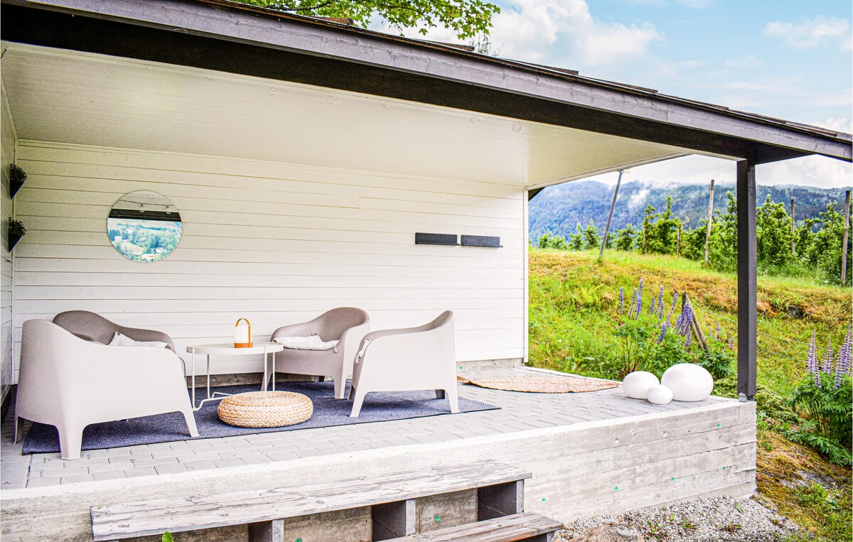 A covered outdoor seating area features two light-colored armchairs and a round table, set on a wooden deck. A circular mirror adorns the wall, while the surrounding landscape showcases lush greenery and distant mountains under a clear sky.