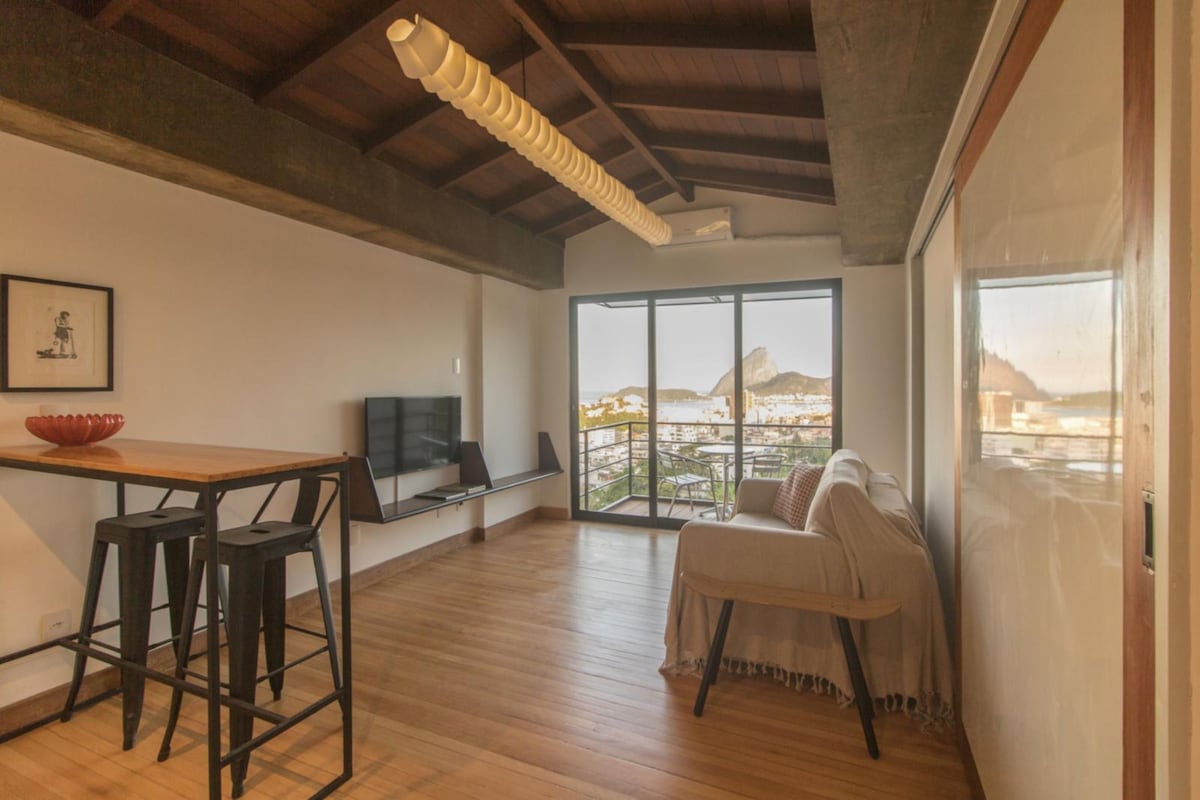 A modern living area features wooden flooring and a high ceiling with exposed beams. A small dining table with four black stools is positioned next to a wall-mounted TV. Large glass doors provide access to a balcony offering a view of the landscape.