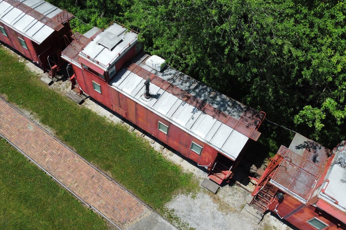 Three red cabooses are arranged in a row along a path surrounded by lush greenery. Each caboose features a flat roof and small windows. The area in front is paved with bricks, extending towards the grassy landscape, creating a welcoming outdoor space.