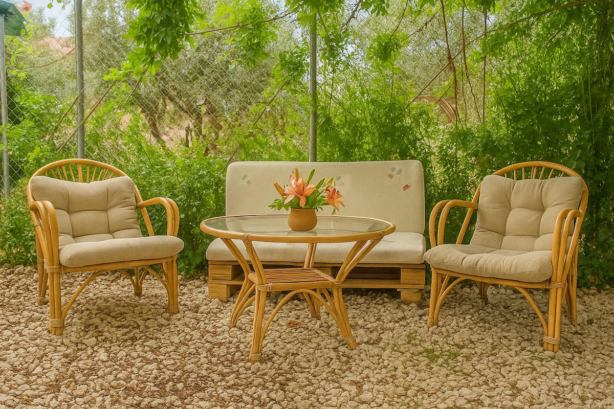 An inviting outdoor seating area features a bamboo sofa and two matching chairs, all adorned with plush cushions. A glass table, centered with a vase of fresh flowers, enhances the tranquil garden setting, surrounded by lush greenery and natural stone flooring.
